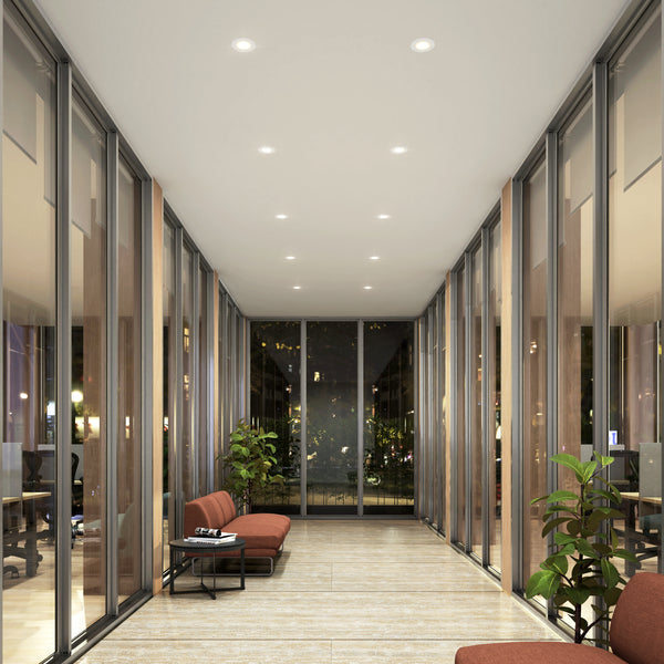 A modern hallway features Skyview 4in Recessed Downlight Slim Panel 5CCT LEDs, floor-to-ceiling glass windows, tan tile floors, red cushioned benches, side tables, and green potted plants. City lights glow outside at night.
