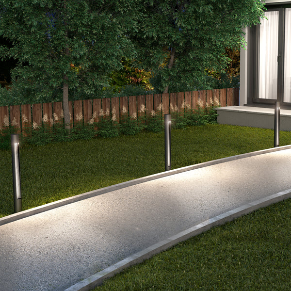 At night, a curved outdoor path is lit by Dark Sky Pro LED Outdoor Pathway Light Sticks. Grass lines the path, with a wooden fence and green trees behind, near a house with large windows.