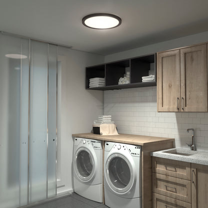 Modern laundry room featuring two front-loading washers, wooden cabinets, open shelves with towels, a countertop, sink, white subway tile backsplash, sliding glass doors, and the Skylight Pro 15" 5K LED Flat Panel Light with adjustable brightness.
