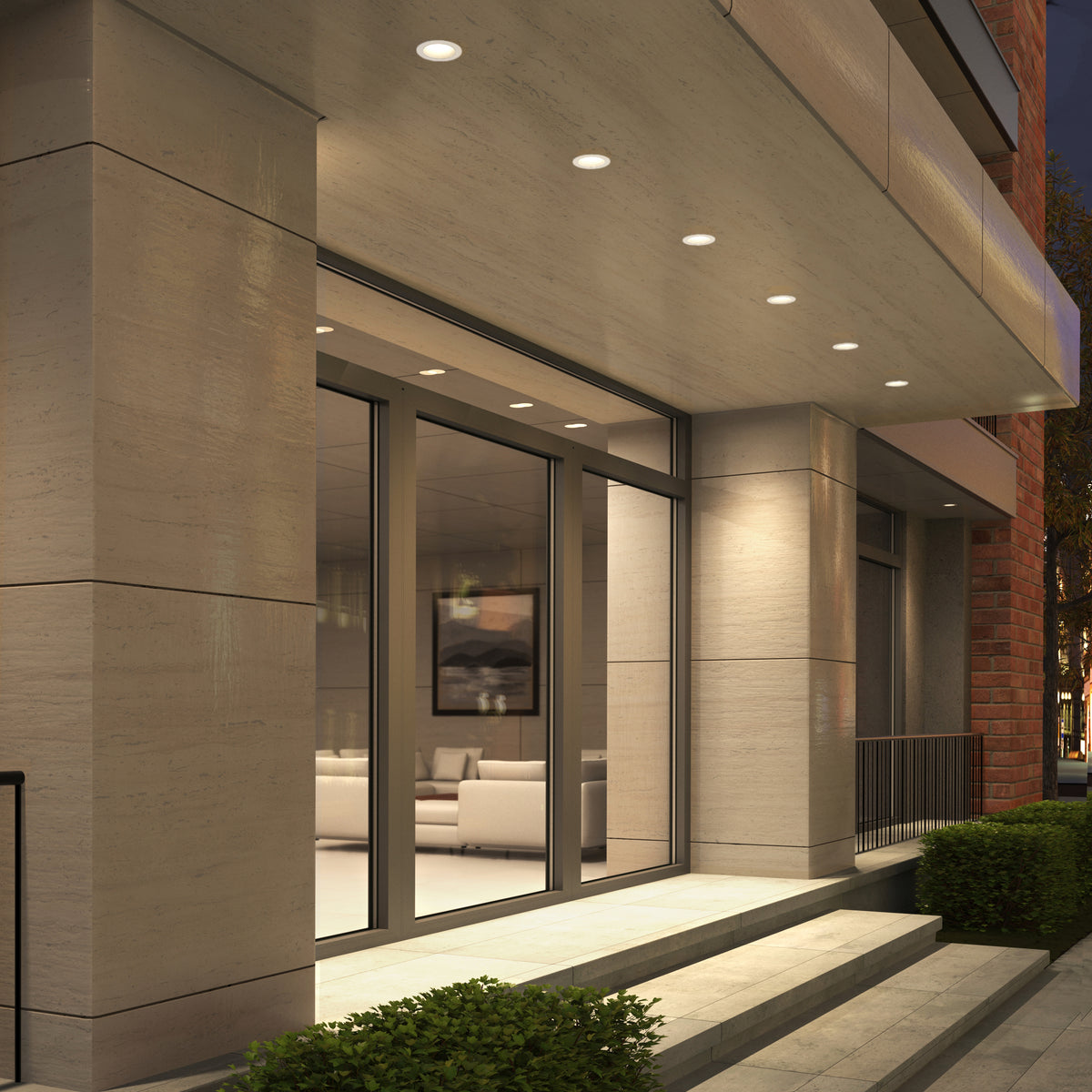 A modern building entrance with large glass doors, wide steps, flanked by green bushes. Skyview 4in Regressed Round, 5CCT LED pot lights with recessed diffusers illuminate the ceiling. Warm indoor light highlights couches inside.
